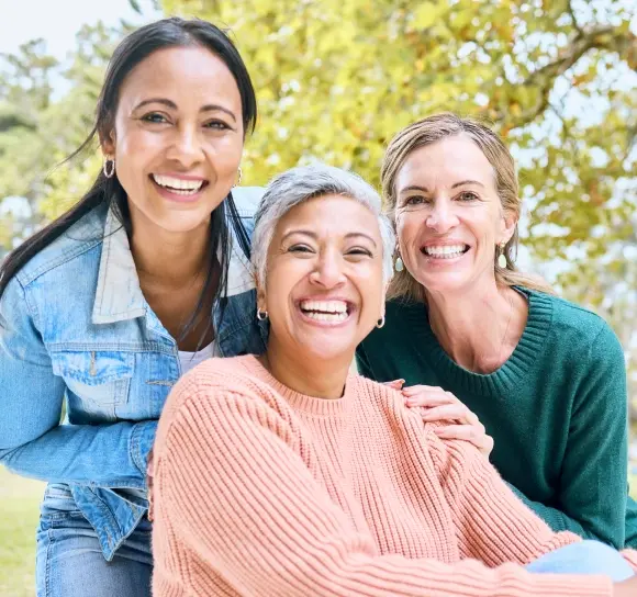 Three smiling women posing together outdoors with trees in background.
